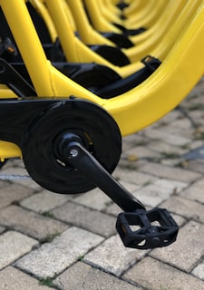 Close-up view of a row of yellow bicycles focused on the pedals and drivetrain components. The bikes are aligned in an orderly manner on a paved surface with light-colored stones.