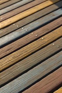 Wooden planks arranged side by side with distinct grain patterns and varying colors, including shades of brown, yellow, and blue. Small metallic screws are visible on the surface.