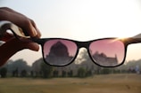 A hand holds up a pair of sunglasses, with the lenses framing a view of a building in the background. The sun shines through the right lens, creating a sunburst effect. The left lens has a slight pink tint. The scene includes trees and a grassy area, suggesting a park or historical site.