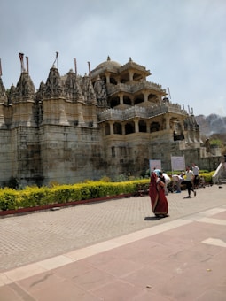 A large, intricately designed stone temple with multiple tiers and ornate carvings. Several people are walking around the temple grounds, with one person wearing a vibrant red garment. The temple is surrounded by a low hedge with yellow flowers, and there is signage near the steps leading up to the structure.