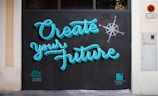 Brightly colored graffiti on a wall reads 'Create your Future' with a compass design nearby. The lettering is in a turquoise blue with a black background, and there are small tags near the bottom by the artists.
