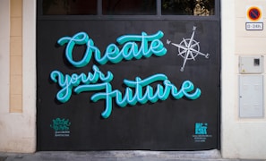 Brightly colored graffiti on a wall reads 'Create your Future' with a compass design nearby. The lettering is in a turquoise blue with a black background, and there are small tags near the bottom by the artists.