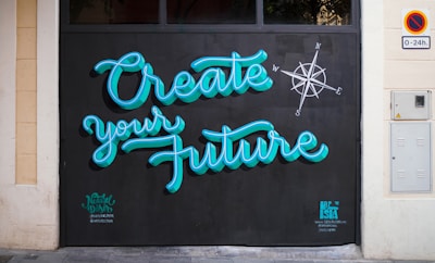 Brightly colored graffiti on a wall reads 'Create your Future' with a compass design nearby. The lettering is in a turquoise blue with a black background, and there are small tags near the bottom by the artists.