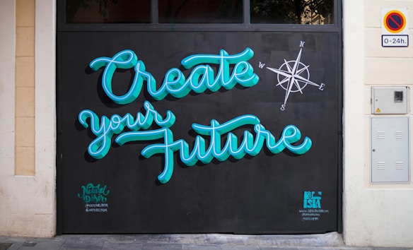 Brightly colored graffiti on a wall reads 'Create your Future' with a compass design nearby. The lettering is in a turquoise blue with a black background, and there are small tags near the bottom by the artists.