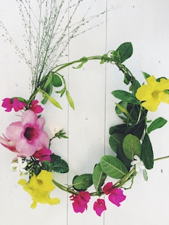 A decorative floral wreath is arranged with a combination of vibrant yellow, pink, and white flowers interspersed with green leaves on a light wooden surface. The circular design is accentuated by sprigs of delicate foliage extending outward.