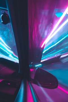 A futuristic scene with vibrant neon lights in shades of pink and blue reflecting off a car's side view mirror and window. The lights create a sense of speed and movement, resembling a tunnel or highway at night.