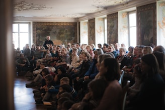 A room filled with a diverse group of people, including both adults and children, seated and attentively facing what appears to be a speaker at the front. The room has ornate decor, with painted walls and windows allowing soft light to filter in.