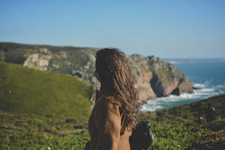 woman looking at cliffs while ocean waves are pounding it during daytime
