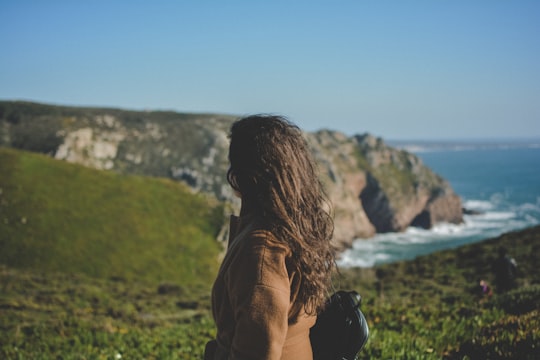 woman looking at cliffs while ocean waves are pounding it during daytime in Sintra-Cascais Natural Park Portugal