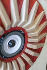 A close-up view of a speaker surrounded by wooden triangular shapes radiating outwards, set against a backdrop of rich, reddish-brown wood.