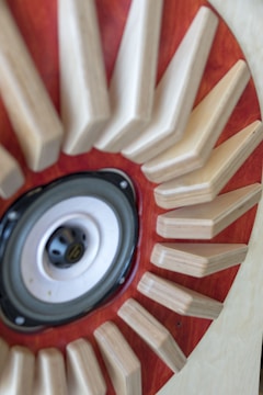 A close-up view of a speaker surrounded by wooden triangular shapes radiating outwards, set against a backdrop of rich, reddish-brown wood.