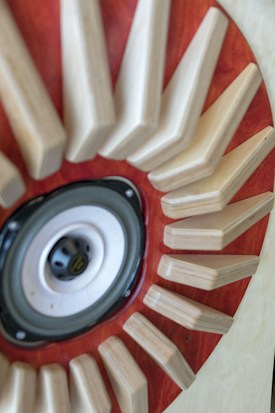 A close-up view of a speaker surrounded by wooden triangular shapes radiating outwards, set against a backdrop of rich, reddish-brown wood.