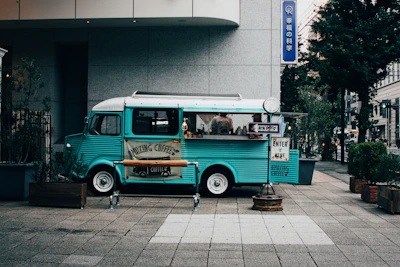 A turquoise vintage coffee van is parked on the sidewalk, surrounded by potted plants and small trees. It has a banner that reads 'Amazing Coffee' displayed on its side. Signs encourage passersby to enter and buy coffee. The setting is urban with modern buildings in the background and a calm street scene.