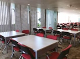 A large, modern conference room is filled with rows of tables and chairs. The chairs have red seats and black backs, and the tables are light-colored. Large windows with vertical blinds allow natural light to fill the space, giving it a bright, airy feel.