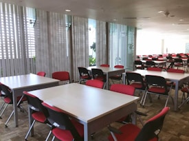 A large, modern conference room is filled with rows of tables and chairs. The chairs have red seats and black backs, and the tables are light-colored. Large windows with vertical blinds allow natural light to fill the space, giving it a bright, airy feel.