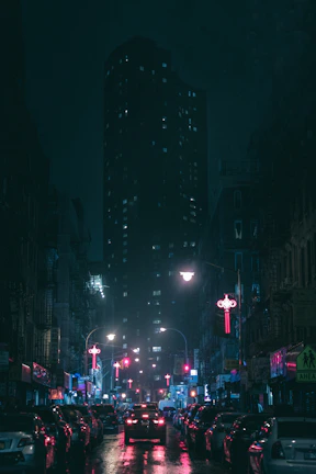 A dark city street with cars lined up, their neon underglow casting colorful reflections on wet pavement.