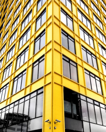 A modern building facade featuring geometric patterns with bright yellow framing and large glass windows. The structure gives a sense of symmetry and order due to its repetitive window design. Two surveillance cameras are visible near the bottom.