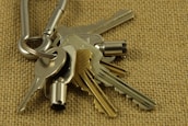 A close-up view of a set of metallic keys attached to a carabiner, placed on a textured burlap surface. The keys vary in shape and size, with some having distinct notches and others appearing more cylindrical.
