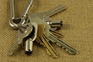 A close-up view of a set of metallic keys attached to a carabiner, placed on a textured burlap surface. The keys vary in shape and size, with some having distinct notches and others appearing more cylindrical.