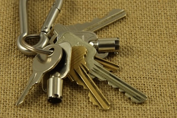 A close-up view of a set of metallic keys attached to a carabiner, placed on a textured burlap surface. The keys vary in shape and size, with some having distinct notches and others appearing more cylindrical.