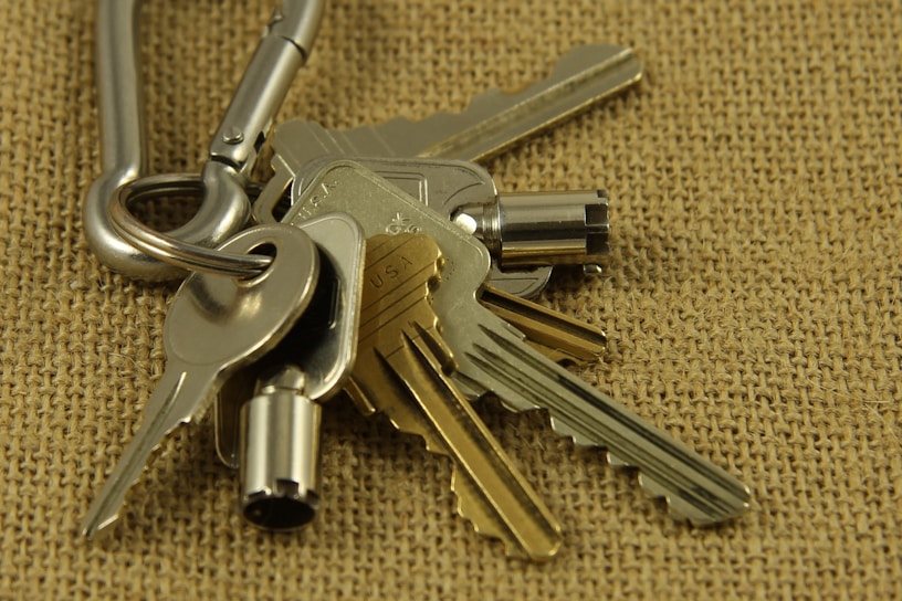 A close-up view of a set of metallic keys attached to a carabiner, placed on a textured burlap surface. The keys vary in shape and size, with some having distinct notches and others appearing more cylindrical.