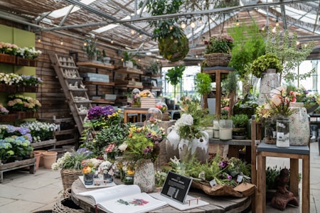 A well-organized indoor garden shop filled with various potted plants and flowers. A large wooden table displays an array of vibrant floral arrangements, ferns, and succulents. There are decorative ceramic pots and vases, a book open to a page on floral design, and a small chalkboard sign with plant information. Wooden shelves and ladders line the walls, holding additional plants and garden ornaments.