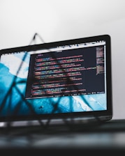 A laptop screen displaying a code editor with multiple lines of colorful code written in a programming language. The background beyond the screen is blurred, emphasizing the screen itself with a clean interface. Various code-related icons and interface details can be seen on the editor.