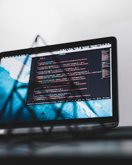A laptop screen displaying a code editor with multiple lines of colorful code written in a programming language. The background beyond the screen is blurred, emphasizing the screen itself with a clean interface. Various code-related icons and interface details can be seen on the editor.