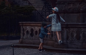 Two children are playing near an ornate stone structure with carved details. One child, wearing a cap and a white dress, is jumping or reaching up while the other child, dressed in a beanie and casual clothes, stands nearby. The scene is set in a dimly lit outdoor area with stairs and wrought iron gates in the background.