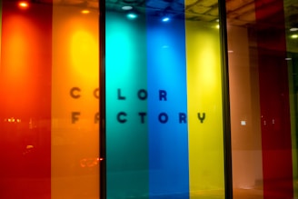 A vibrant display featuring vertical panels of translucent colors, including red, orange, yellow, blue, and green. The text 'COLOR FACTORY' is subtly visible behind the panels, casting a shadow effect.
