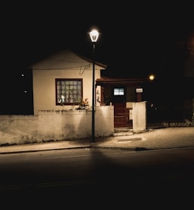 A small house is visible at night, illuminated by a streetlamp. The building is surrounded by a low white wall, and a paved sidewalk leads to its entrance. The warm light from the lamp casts shadows on the ground, adding a tranquil atmosphere.