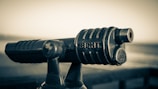 A vintage binocular telescope with the words 'SIGHT' and 'FOCUS' engraved on it, positioned at a scenic viewpoint. The image has a sepia tone, giving it an aged and nostalgic feel. The telescope is slightly worn, indicating frequent use or age. In the background, a blurred landscape suggests an open, possibly coastal or mountainous, setting.