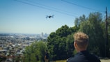A person is looking at a drone flying over a cityscape. The scene is set on a hilltop with urban sprawl visible in the distance. Trees and greenery are in the foreground, blending with the blue sky above.