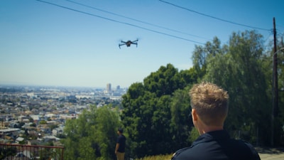 A person is looking at a drone flying over a cityscape. The scene is set on a hilltop with urban sprawl visible in the distance. Trees and greenery are in the foreground, blending with the blue sky above.