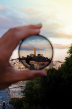 A foreground hand holds a circular lens, magnifying an urban landscape with a prominent tower surrounded by trees and buildings. The surrounding scene is slightly blurred, with greenery on the right and a city street extending into the distance on the left. The sky is partly cloudy with a warm gradient of colors.