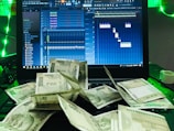 A laptop displays a digital audio workstation interface, possibly for music production, with multiple audio tracks and controls visible on the screen. In front of the laptop, there is a scattered pile of currency notes, predominantly Indian rupees. The scene is accented with green lighting.