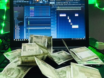 A laptop displays a digital audio workstation interface, possibly for music production, with multiple audio tracks and controls visible on the screen. In front of the laptop, there is a scattered pile of currency notes, predominantly Indian rupees. The scene is accented with green lighting.