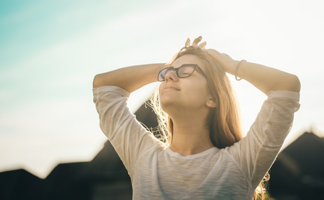 Uma mulher em um dia de sol com os braços recostados em cima da cabeça e os olhos fechados, expressando sentimento de paz