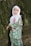 A young child in a colorful, floral-patterned outfit stands beside a tree. The child's headscarf features intricate floral designs and soft pastels, contrasting with the vibrant colors of the outfit. The backdrop consists of greenery, with long drooping branches adding texture to the scene.