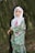 A young child in a colorful, floral-patterned outfit stands beside a tree. The child's headscarf features intricate floral designs and soft pastels, contrasting with the vibrant colors of the outfit. The backdrop consists of greenery, with long drooping branches adding texture to the scene.