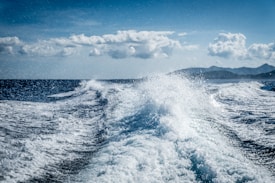 Waves churn in the ocean, creating a frothy white wake against a backdrop of deep blue water. The sky above is partly cloudy, casting soft shadows over the distant mountainous coastline.