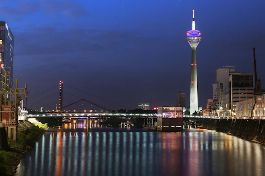 A nighttime cityscape featuring a brightly illuminated telecommunications tower beside a river. The tower, partially lit with red and purple lights, stands out against a deep blue sky. Below, the river reflects the colorful lights from the nearby structures and bridge. Modern buildings and cranes are visible along the riverbank.