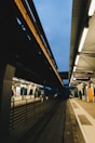 A modern train station platform featuring overhead tracks with a partially visible sky. Fluorescent lighting illuminates the station, highlighting the sleek architectural lines. The platform is empty, with a few visible objects such as signs, information displays, and railings.