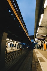A modern train station platform featuring overhead tracks with a partially visible sky. Fluorescent lighting illuminates the station, highlighting the sleek architectural lines. The platform is empty, with a few visible objects such as signs, information displays, and railings.