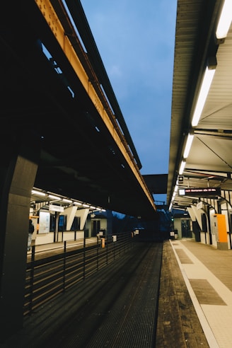 A modern train station platform featuring overhead tracks with a partially visible sky. Fluorescent lighting illuminates the station, highlighting the sleek architectural lines. The platform is empty, with a few visible objects such as signs, information displays, and railings.