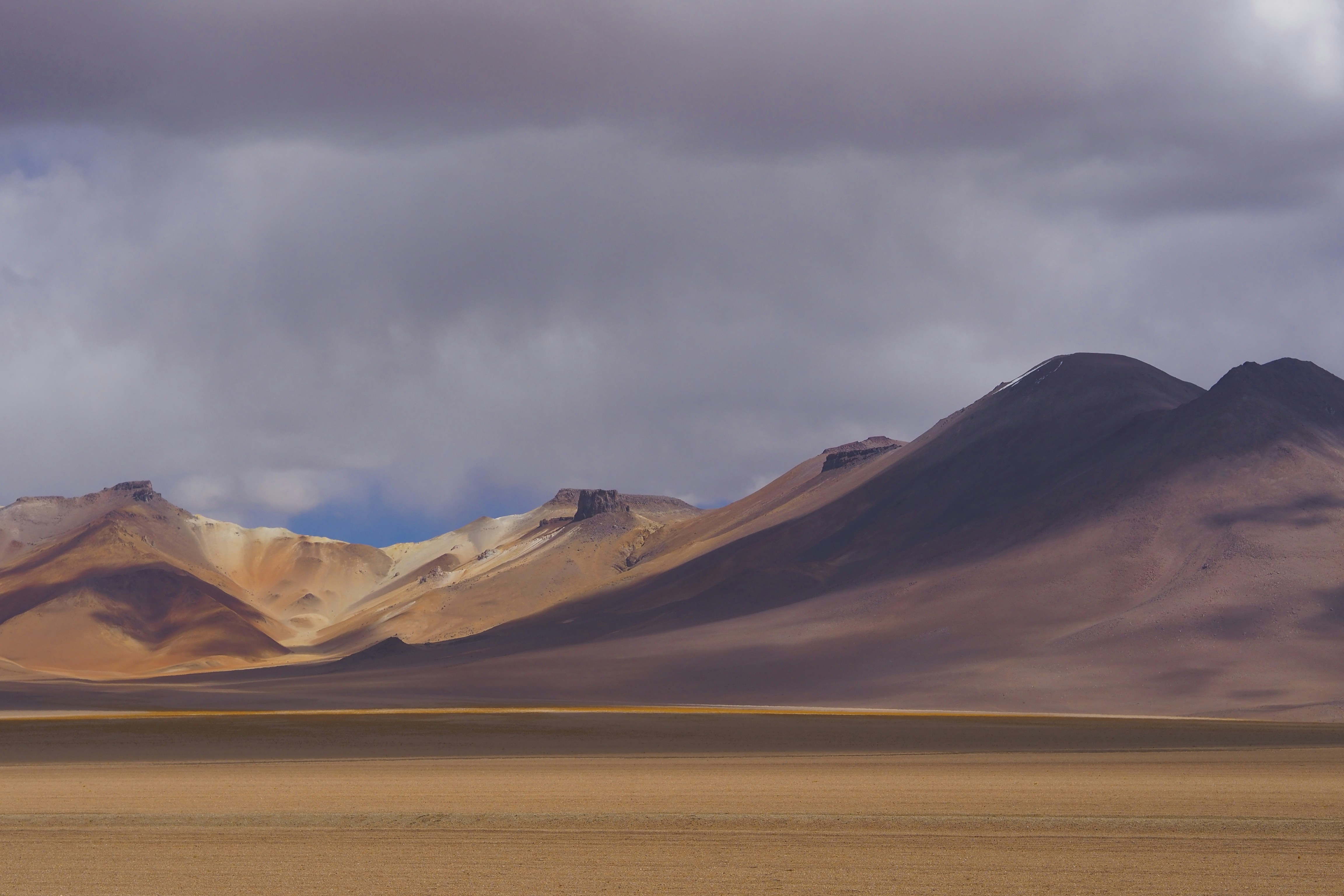 a group of mountains in the distance under a cloudy sky, A scene from a small desert valley on the way to the Salar de Uyuni in Bolivia.