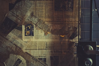A collage of old, yellowed newspaper clippings is arranged haphazardly, with a visible sports article featuring a Lakers jersey number 8. The papers look worn and slightly tattered, creating a vintage or nostalgic feel. The setting seems to be indoors, with a metal door or window frame partially visible on the right.