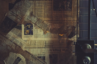 A collage of old, yellowed newspaper clippings is arranged haphazardly, with a visible sports article featuring a Lakers jersey number 8. The papers look worn and slightly tattered, creating a vintage or nostalgic feel. The setting seems to be indoors, with a metal door or window frame partially visible on the right.