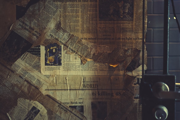 A collage of old, yellowed newspaper clippings is arranged haphazardly, with a visible sports article featuring a Lakers jersey number 8. The papers look worn and slightly tattered, creating a vintage or nostalgic feel. The setting seems to be indoors, with a metal door or window frame partially visible on the right.