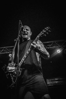 A musician passionately performs on stage, gripping an electric guitar and singing into a microphone. The scene is energized, captured in black and white, highlighting the intensity of the performance. Tattoos are visible on the musician's arms, and stage lights illuminate him from behind, adding to the dramatic atmosphere.
